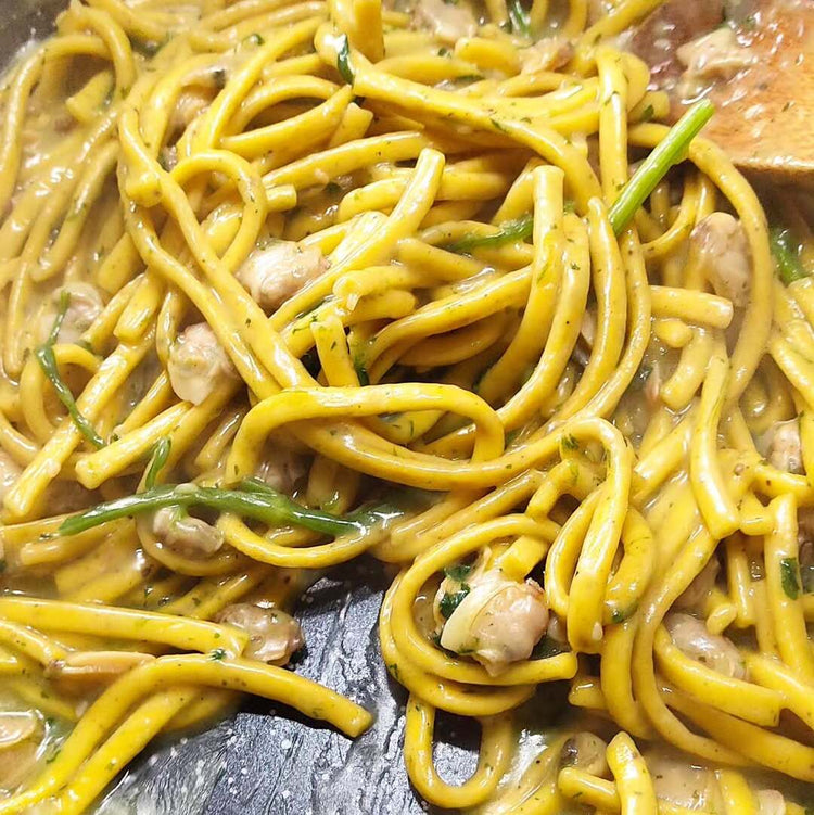 Eine Nahaufnahme zeigt Linguine Limone e Pepe, die in einer Pfanne cremig mit frischen Muscheln und einem aromatischen Sud vermengt werden
