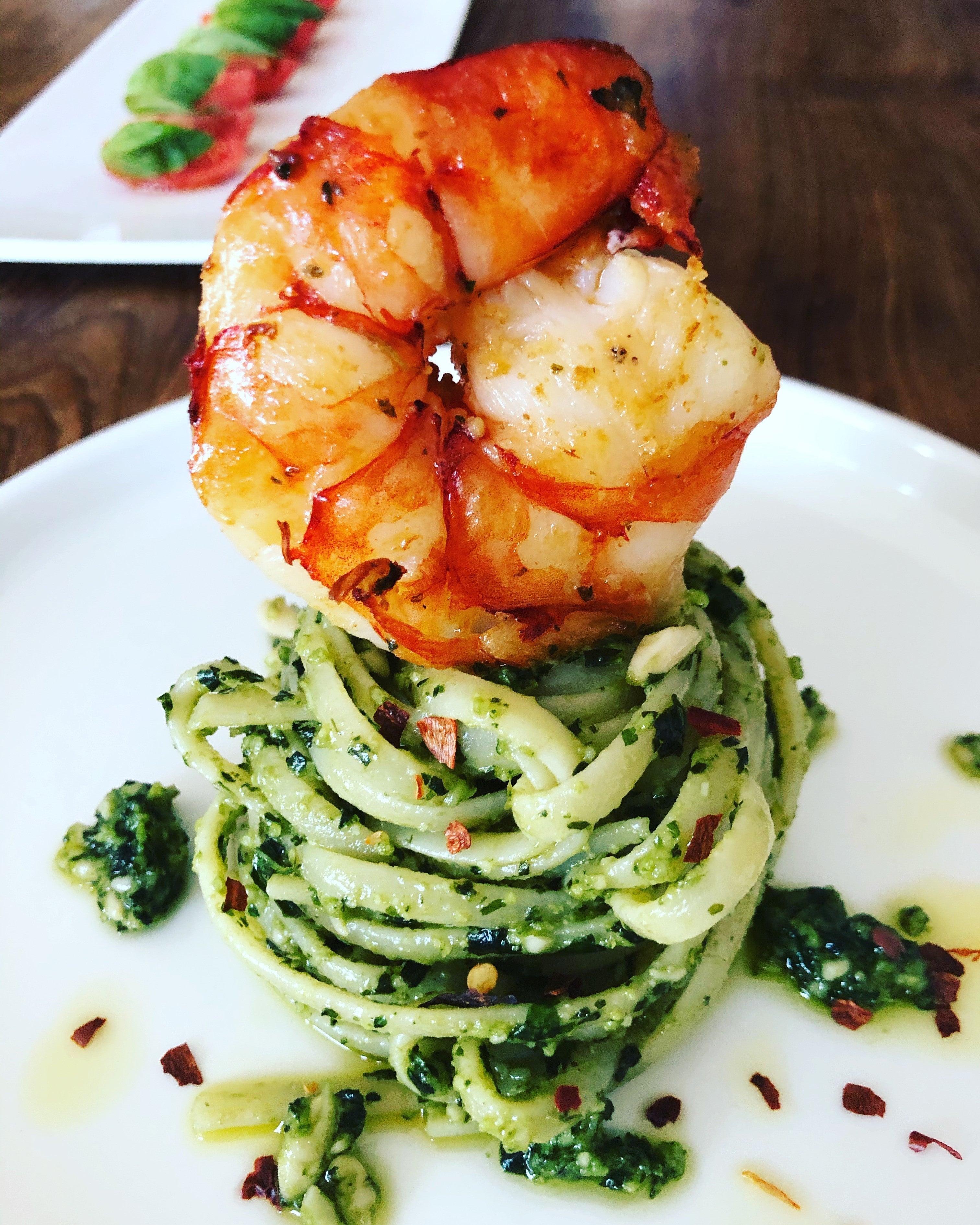 Elegant angerichtete Linguine mit grünem Basilikum-Pesto, spiralförmig als Nest auf einem weißen Teller aufgetürmt, getoppt mit einer gebratenen Wildfanggarnele; garniert mit Chili-Flocken.