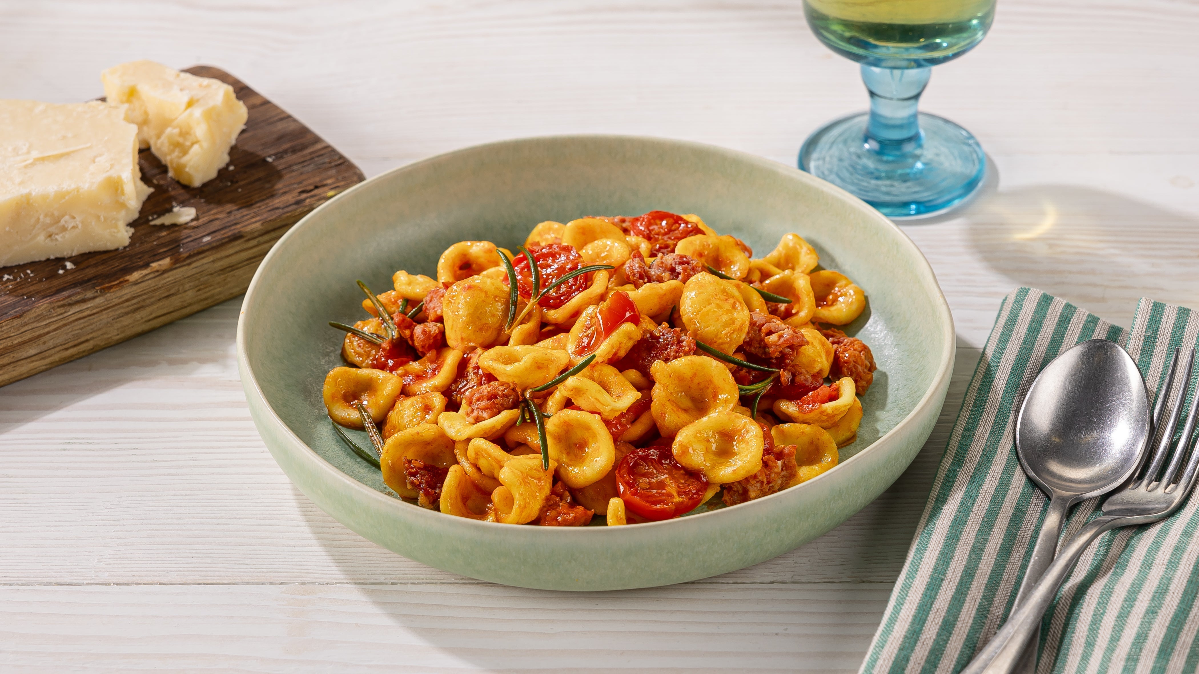 Orecchiette mit Tomatensauce in grüner Schale, daneben Parmesanstück, Weinglas und Besteck auf gestreifter Serviette