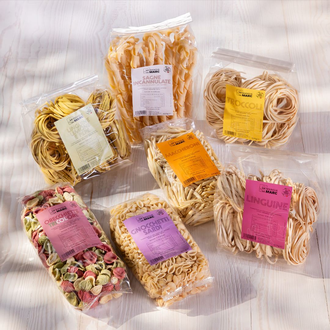 Sechs verschiedene Sorten frische Pasta in transparenten Verpackungen mit bunten Etiketten, darunter Nudelnester, Tagliatelle und bunte Pasta mit Gemüsezusätzen, auf Holzuntergrund