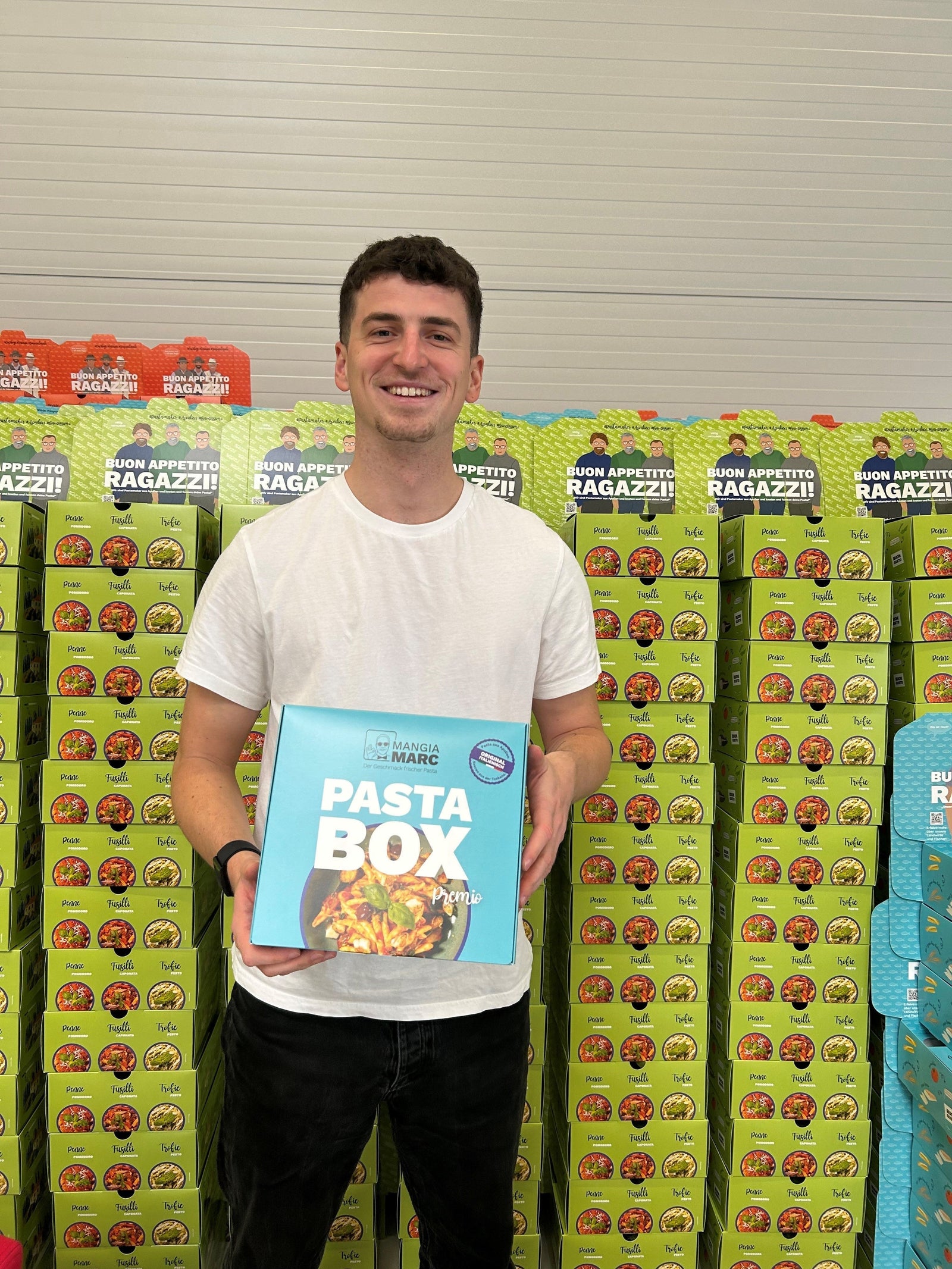 Mann hält eine "Pasta Box"-Verpackung in den Händen, im Hintergrund gestapelte Kartons in einem Lager