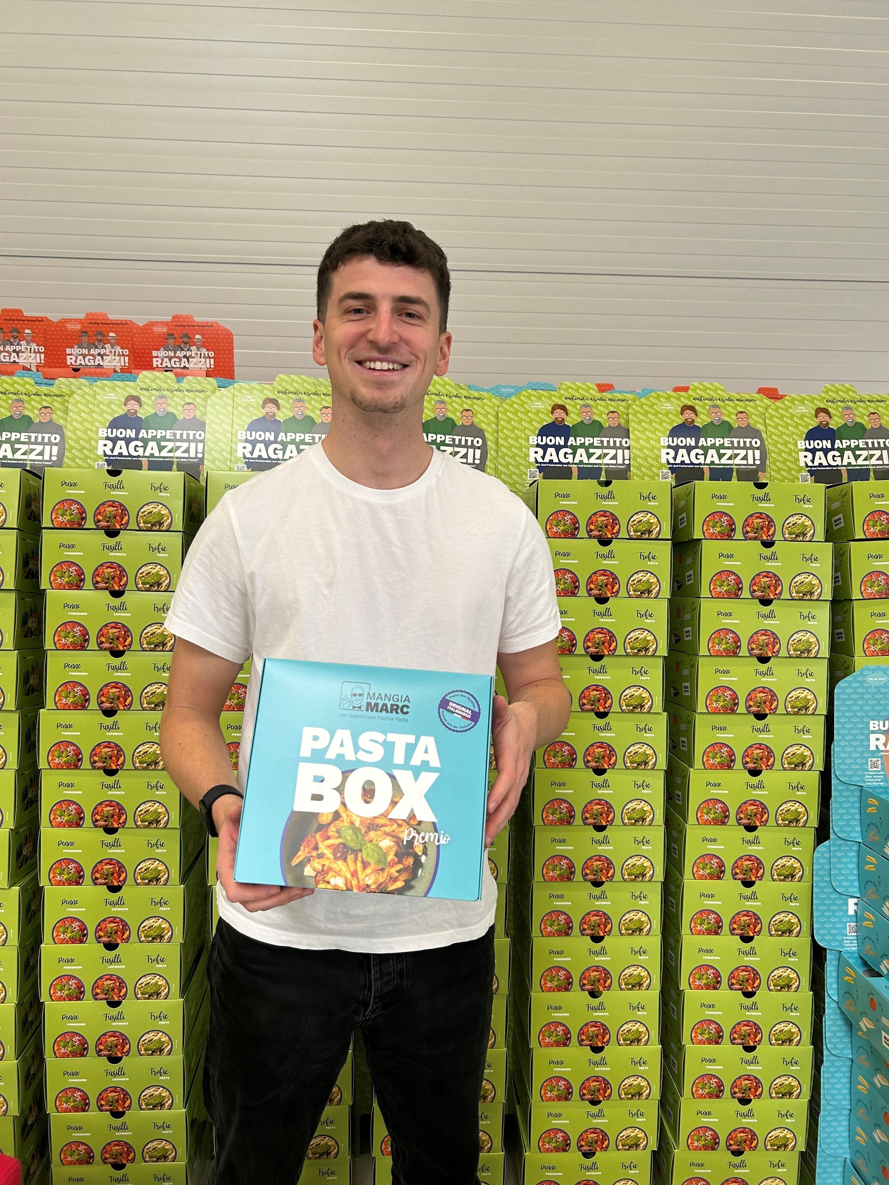 Mann hält eine "Pasta Box"-Verpackung in den Händen, im Hintergrund gestapelte Kartons in einem Lager