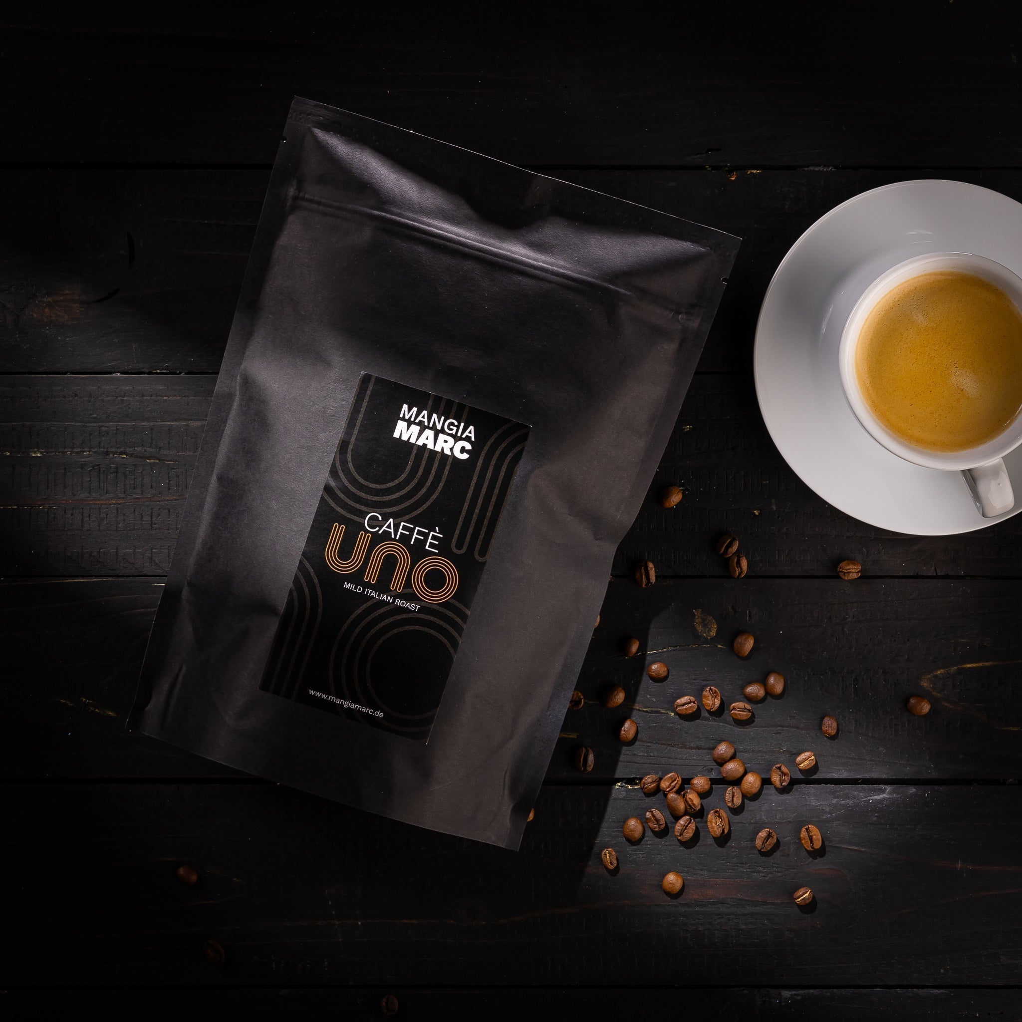 Verpackung "Mamma Marc Caffè Uno" neben einer Tasse Espresso und verstreuten Kaffeebohnen auf dunklem Holztisch